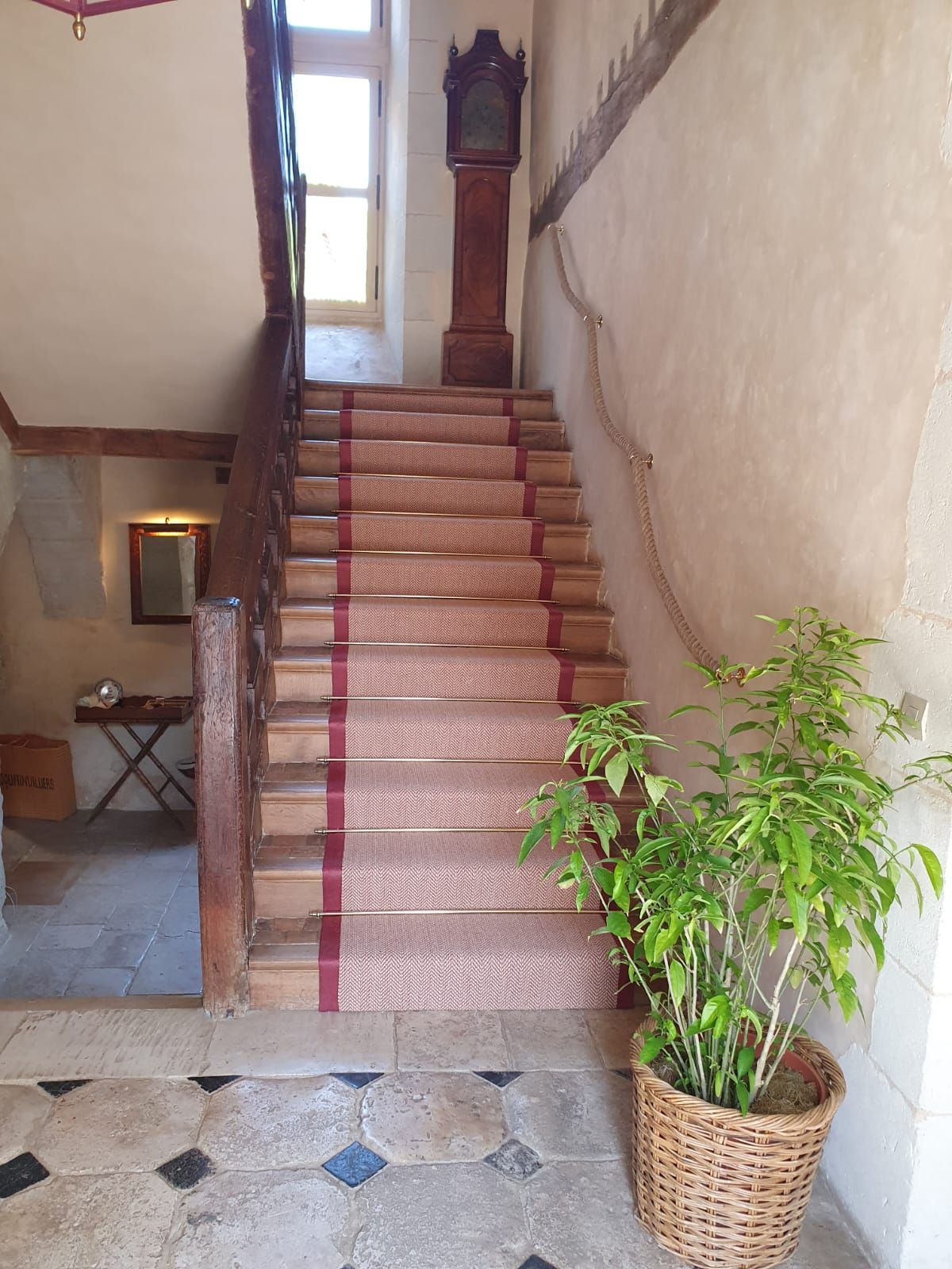Un escalier avec un tapis rouge. Une horloge de parquet trône au sommet. Une plante en pot se trouve à droite.