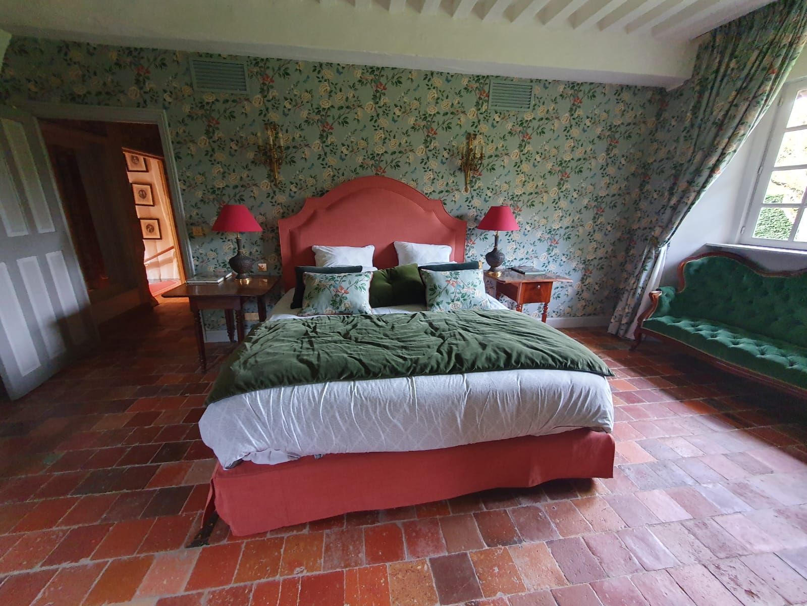 Chambre avec cadre de lit rouge, literie verte, papier peint à motifs et un petit canapé vert.
