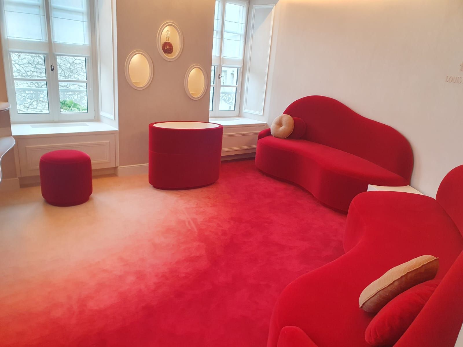 Un canapé rouge aux formes originales et son pouf assorti dans une pièce à la moquette rouge. On aperçoit des fenêtres aux cadres blancs et aux petites décorations murales.