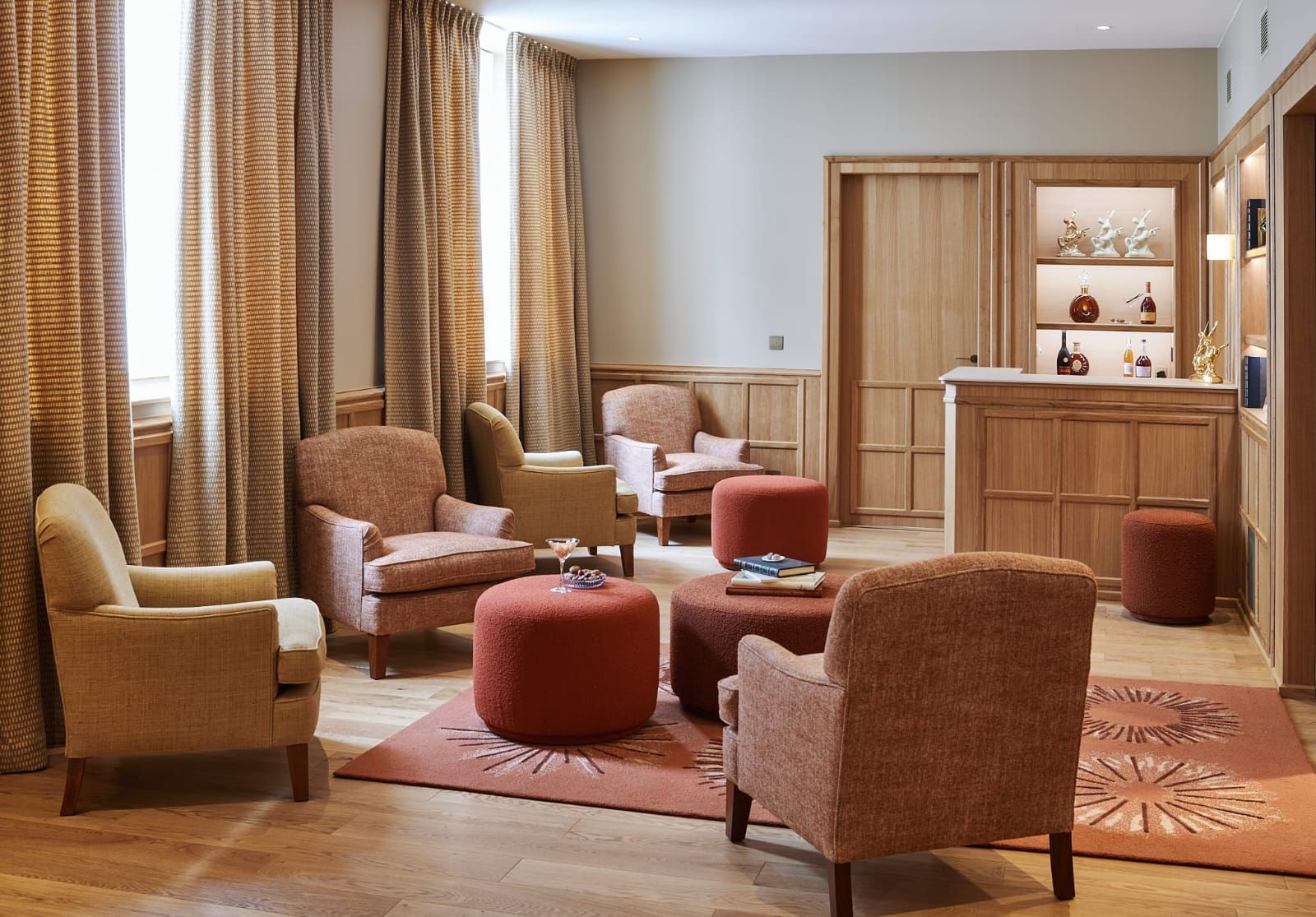 Espace salon confortable avec fauteuils et poufs rembourrés dans des tons pêche, beige et marron, avec des accents de bois clair et un petit bar.