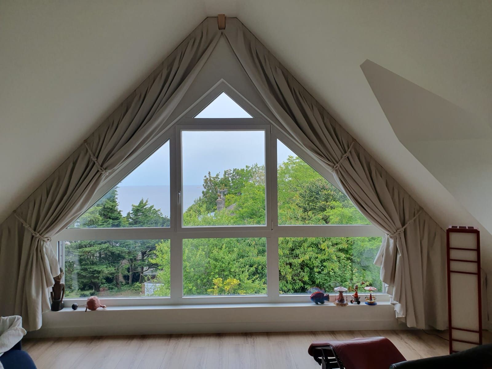 Une fenêtre en angle avec des moulures blanches et des rideaux légers donne sur des arbres verdoyants. La pièce est dotée d'un plafond mansardé et de parquet.