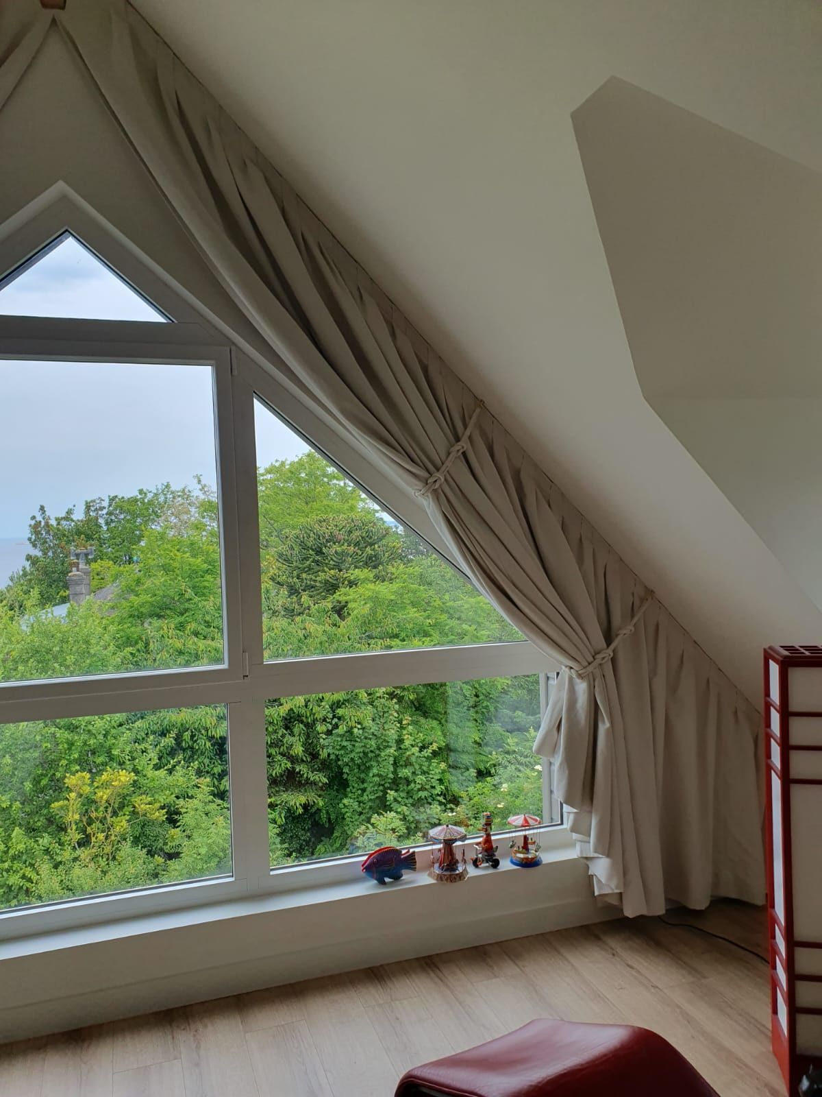 Vue intérieure d'une lucarne avec des rideaux couleur crème. La fenêtre offre une vue sur des arbres verdoyants et un ciel bleu.