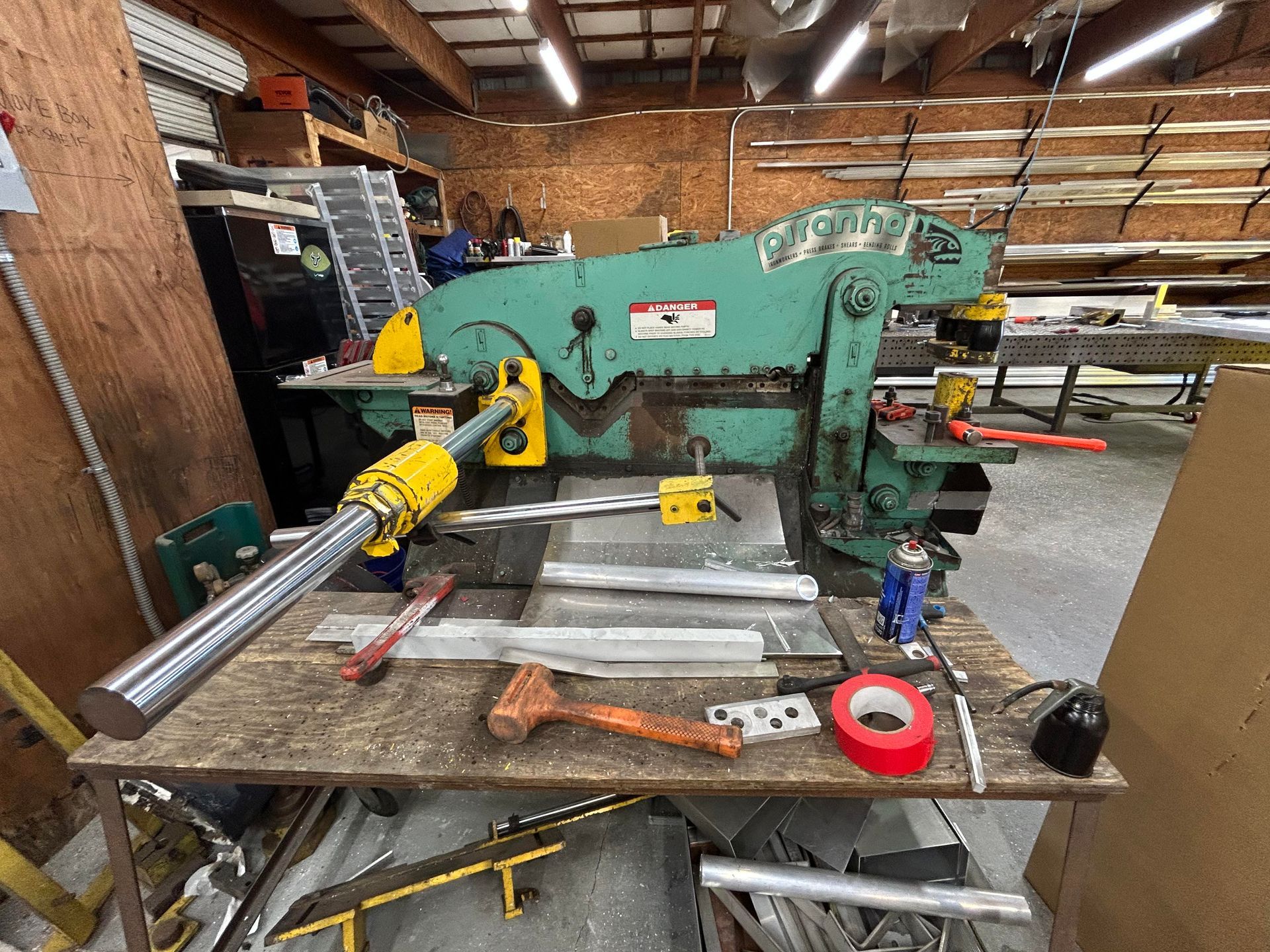 A large green metal shear machine on a workbench in a workshop. Tools and materials are scattered.