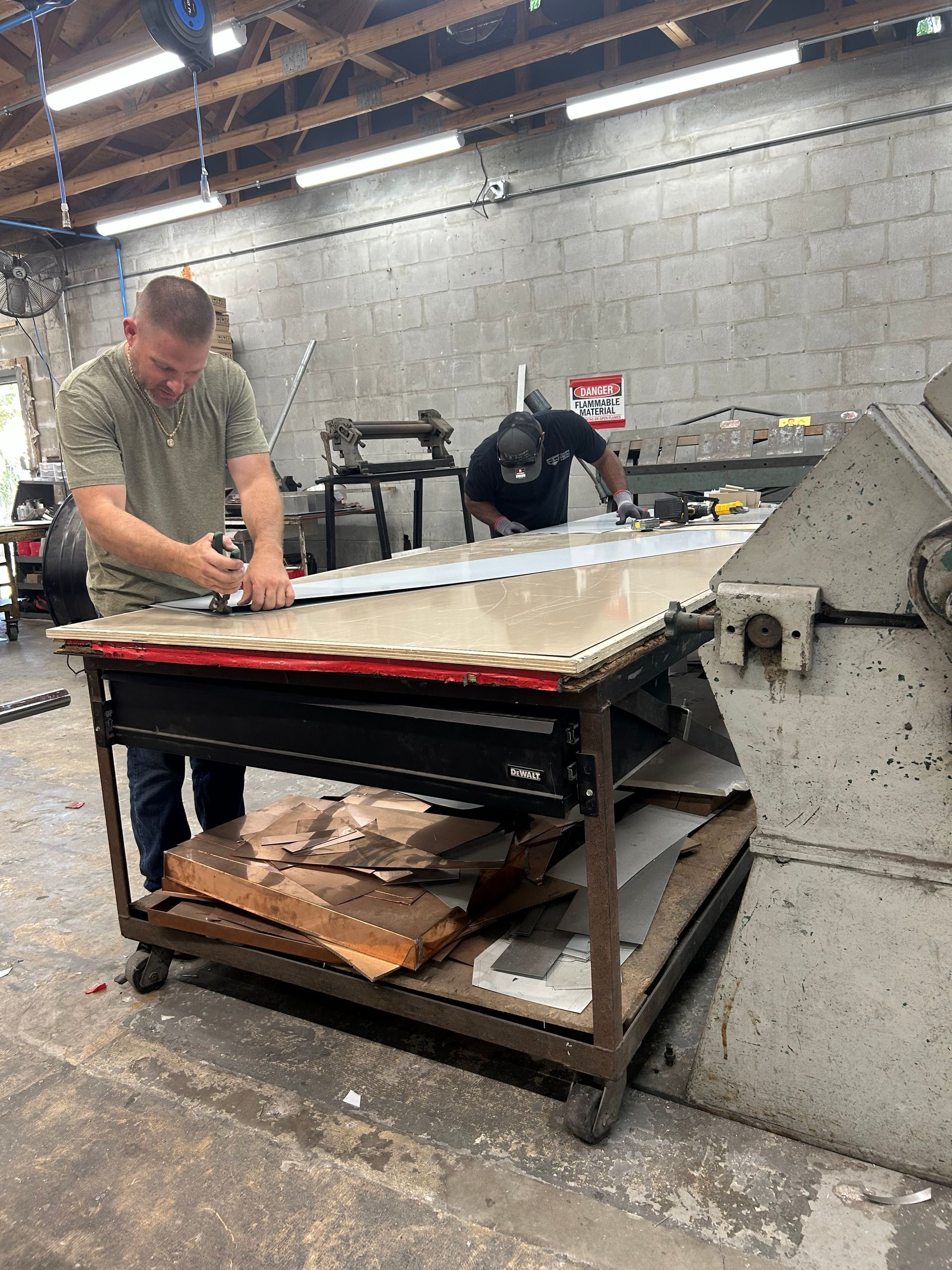 Two men working with metal sheets in a workshop. One cuts with a tool, the other works in the background.