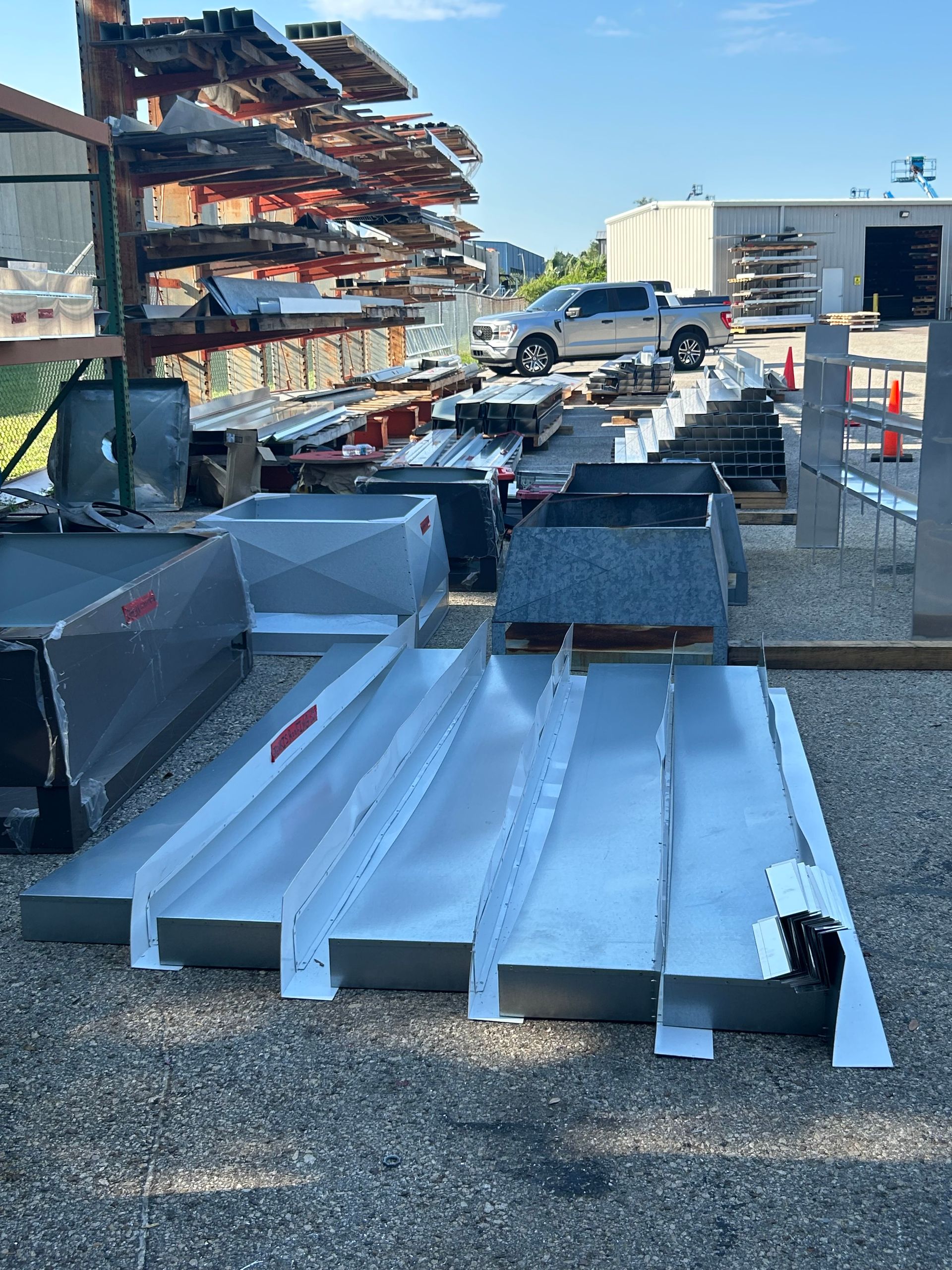 Metal roofing components laid out on gravel with a truck and storage racks in the background.