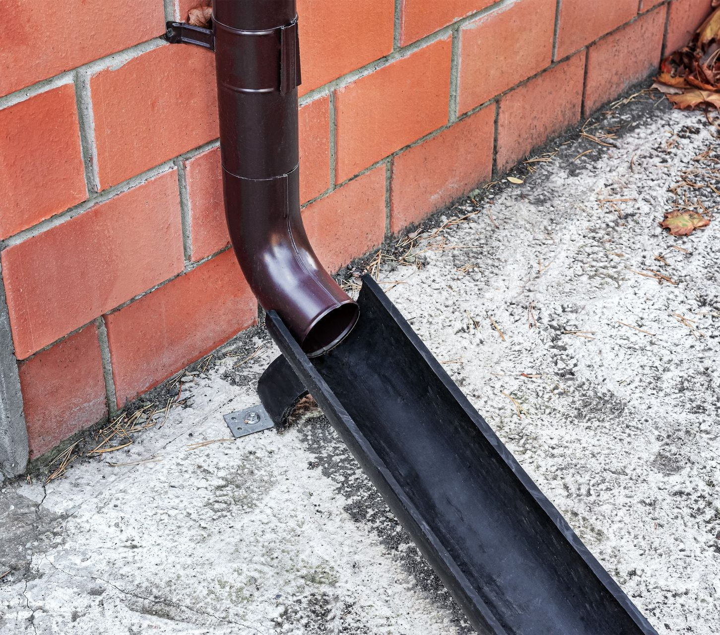Brown gutter downspout emptying into a black trough on a concrete surface near a red brick wall.