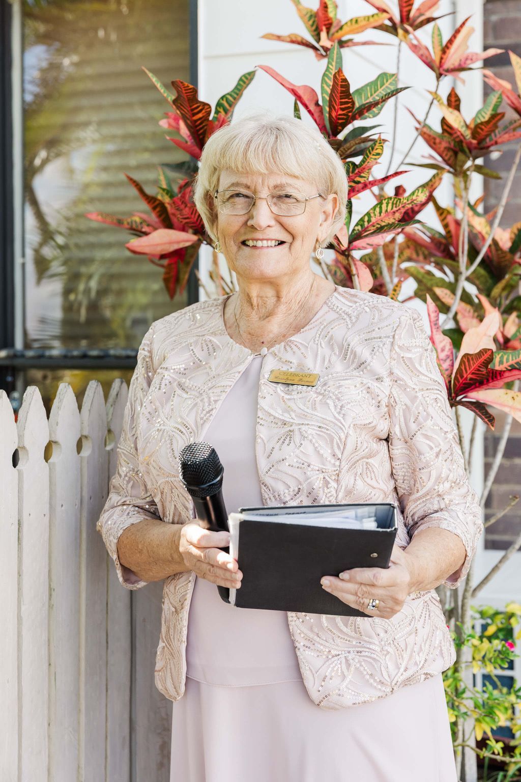 Smiling woman holding a microphone and tablet, standing outdoors beside red-leaf plants and a white fence. — Carolynn Farmer-Celebrant In Emu Park, QLD