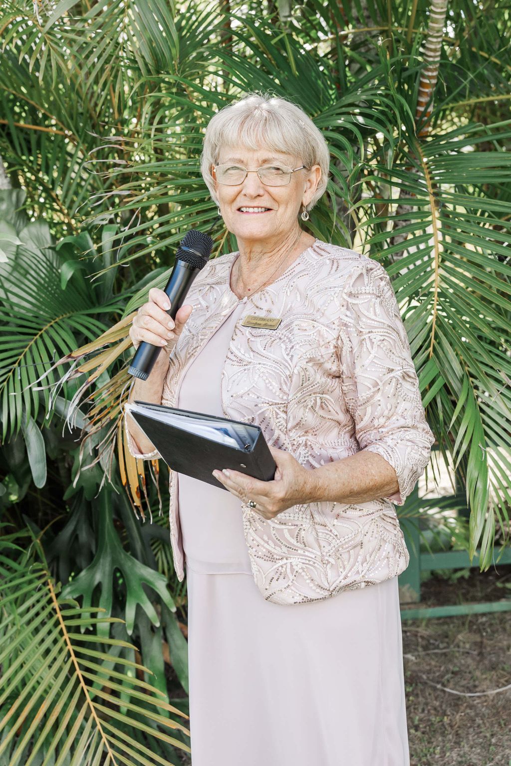 Woman speaking into a microphone while holding a folder, standing in front of lush green plants. — Carolynn Farmer-Celebrant In Emu Park, QLD