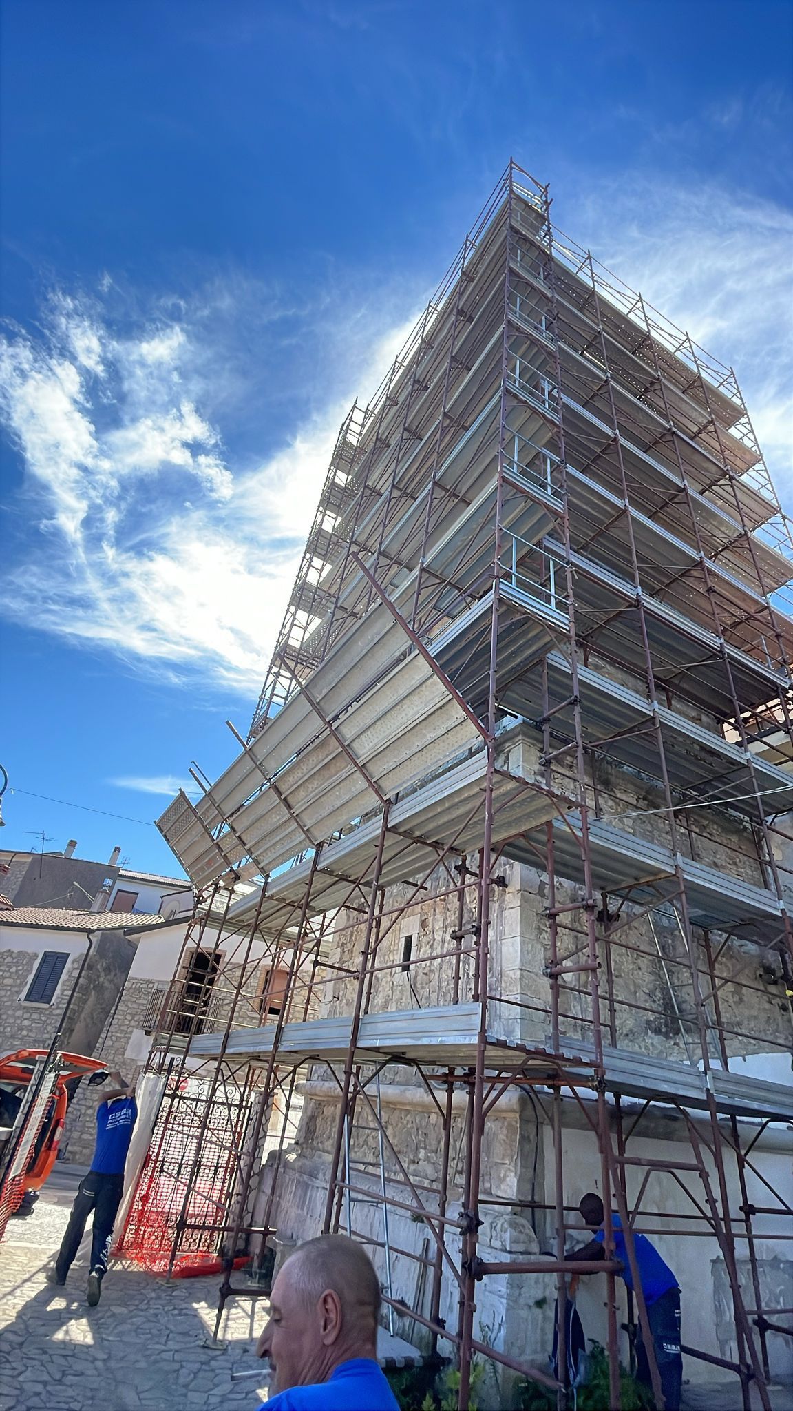 Edificio in ristrutturazione, coperto da impalcature, con operai. Cielo azzurro brillante.