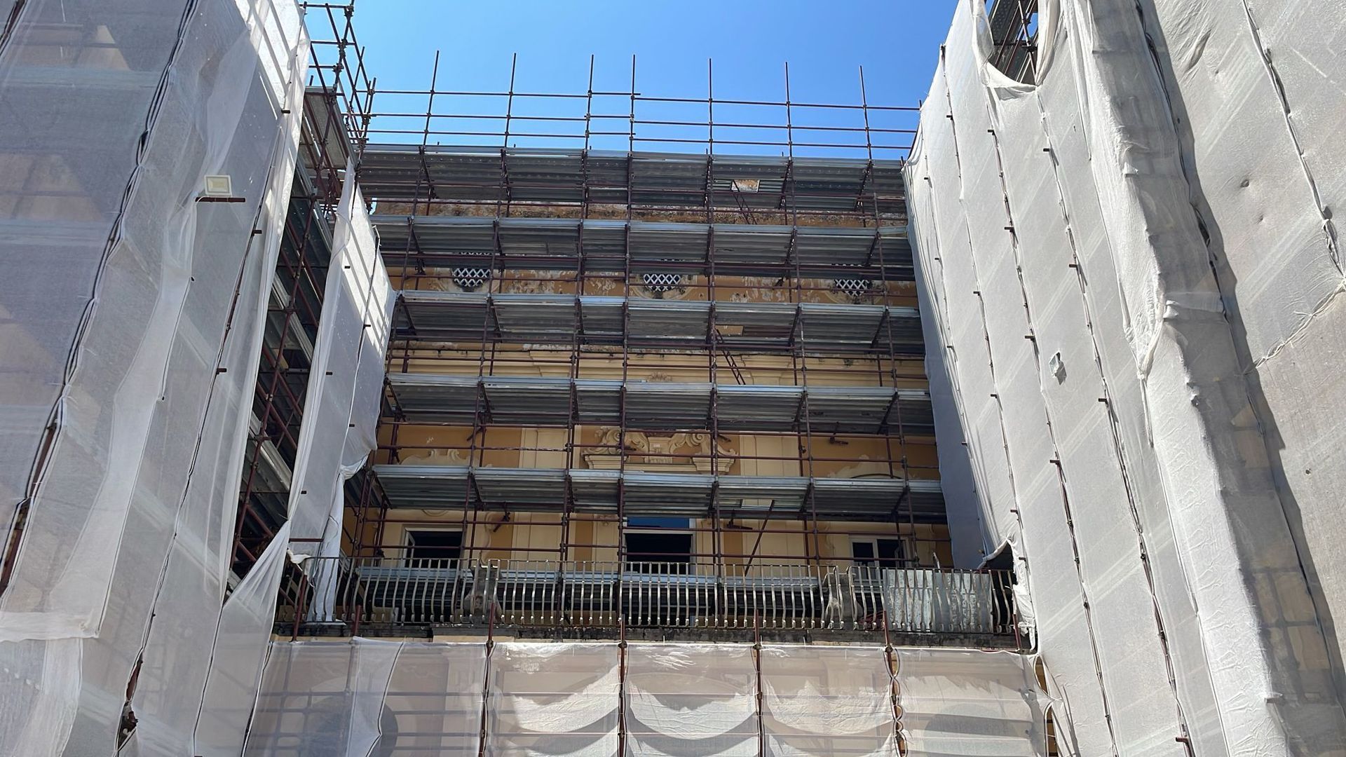 Facciata di un edificio in costruzione, coperta da impalcature e reti protettive, contro un cielo azzurro e terso.