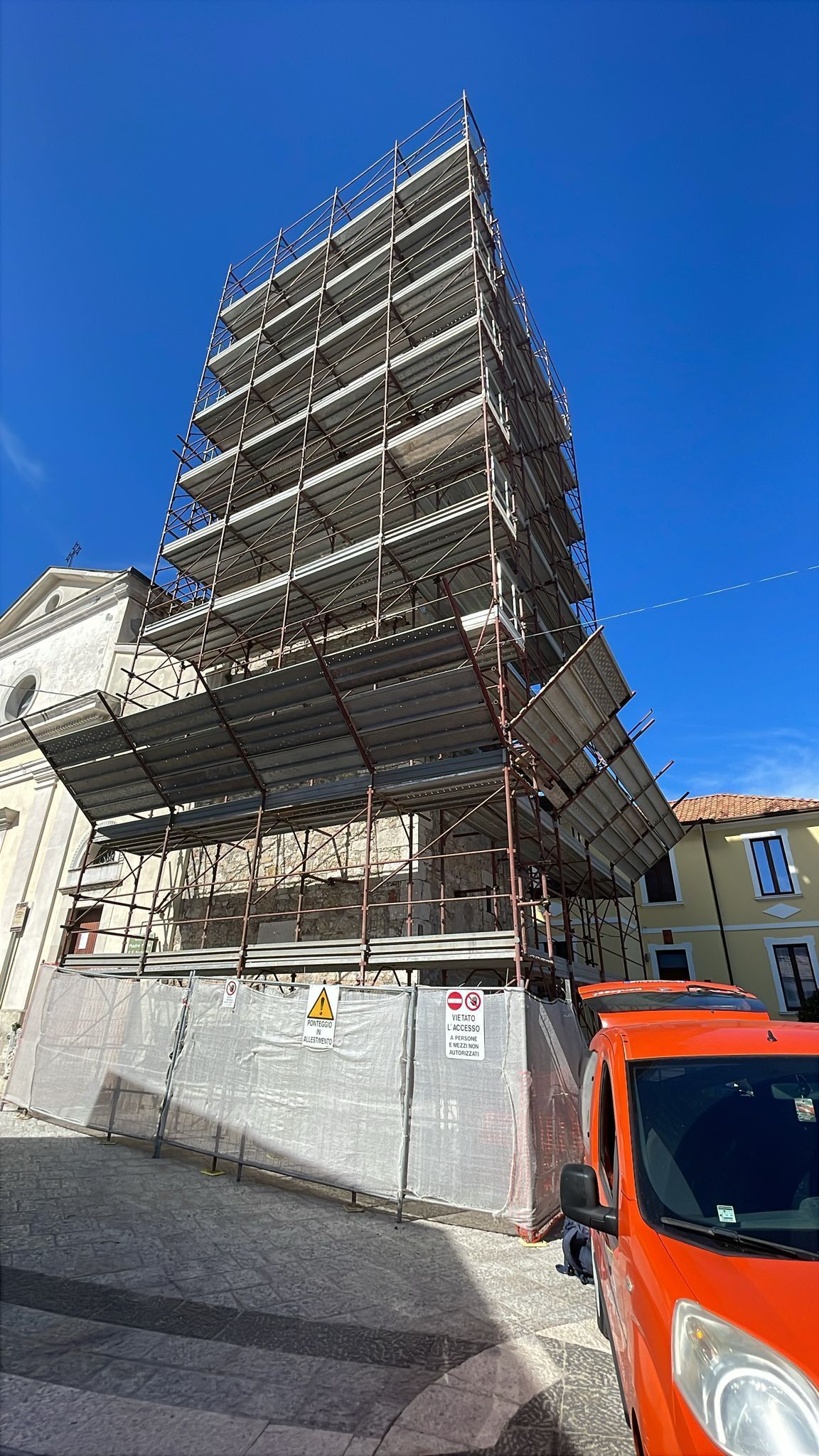 Un'impalcatura circonda l'angolo di un edificio. Un furgone arancione parcheggiato sulla destra. Cielo azzurro.