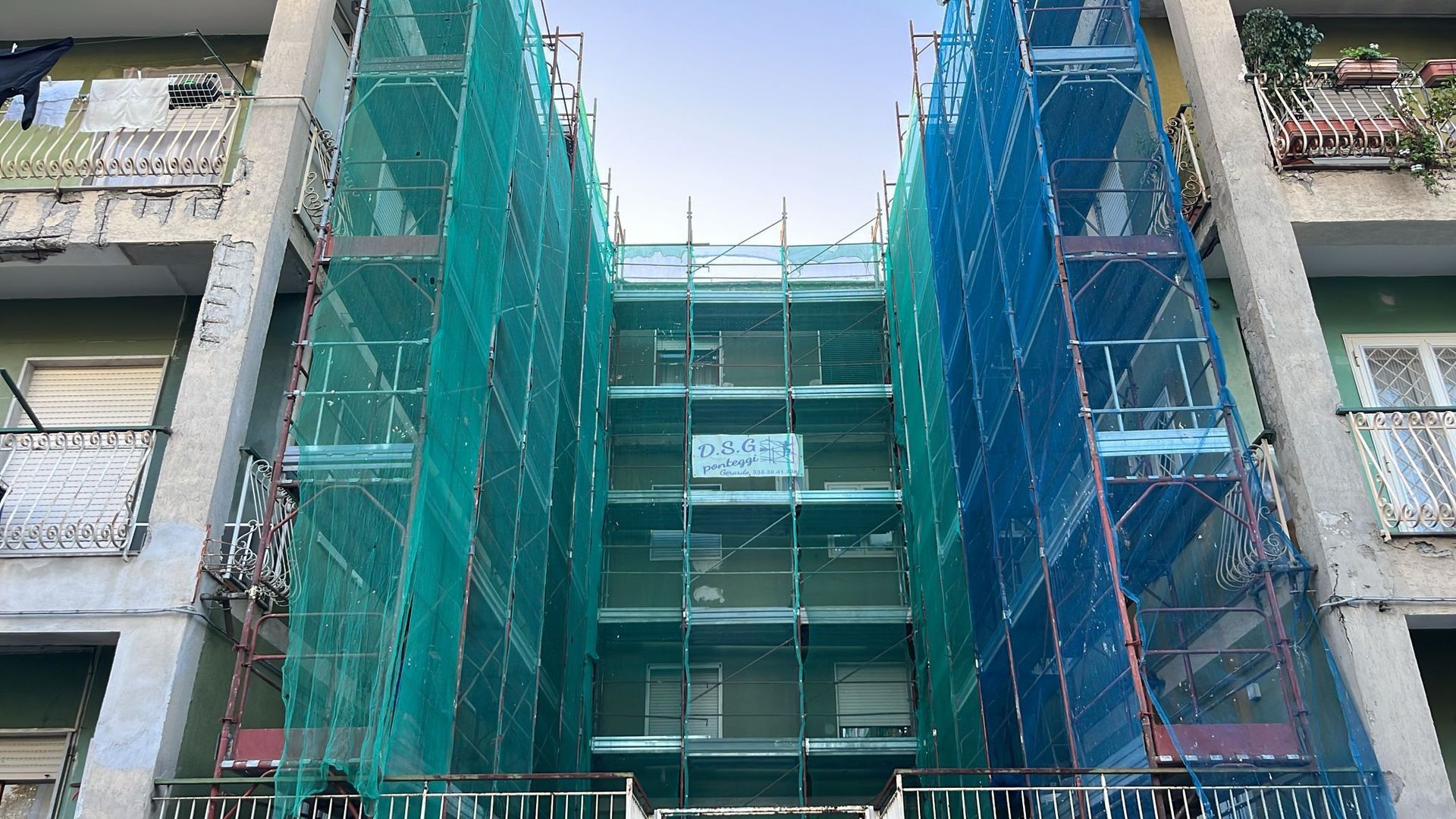 Un'impalcatura con rete di sicurezza verde e blu ricopre la facciata di un edificio, probabilmente in fase di ristrutturazione.