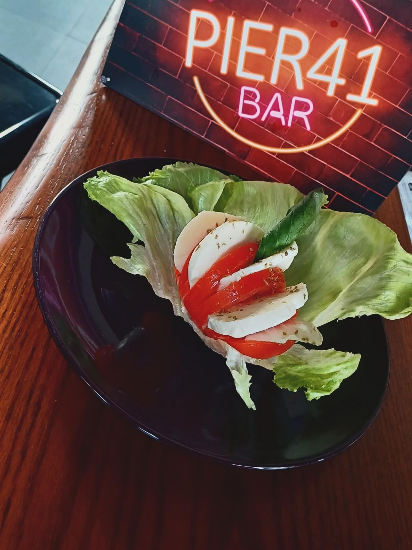 Insalata Caprese in una ciotola nera su un tavolo di legno, con l'insegna luminosa del PIER 41 BAR sullo sfondo.