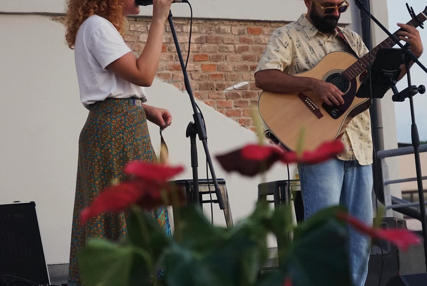 Due musicisti si esibiscono all'aperto, uno canta al microfono e l'altro suona la chitarra, con dei fiori in primo piano.