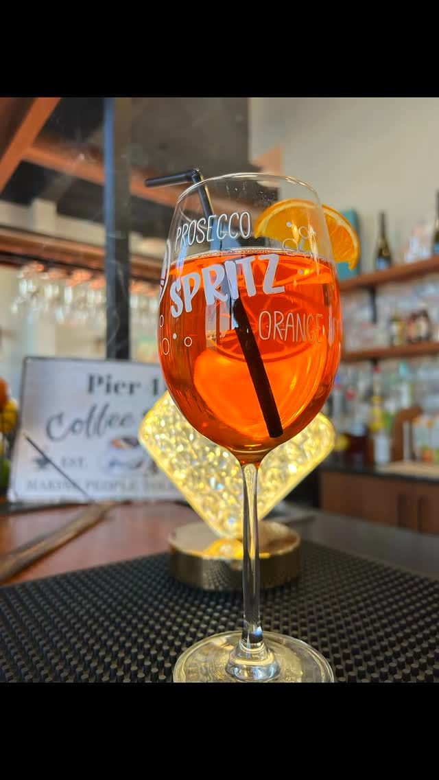 Un Orange Spritz in un bicchiere con stelo su un tavolino da caffè con scaffali sfocati e un'insegna sullo sfondo.