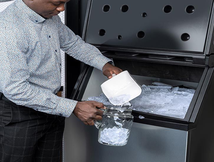 Man in a dress shirt scooping ice from a machine into a pitcher. Black machine, ice, bright setting.
