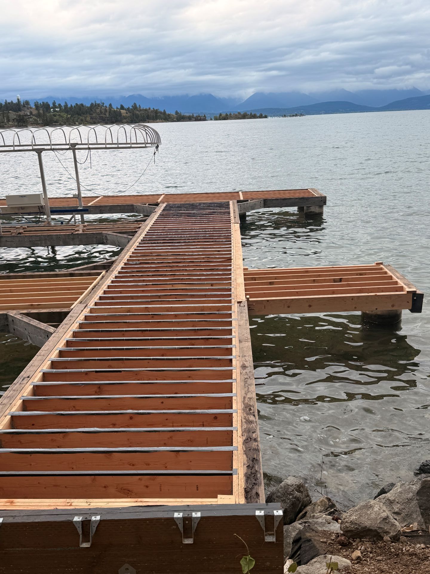 Wooden dock extending into a calm lake under a cloudy sky, with distant mountains and shoreline.