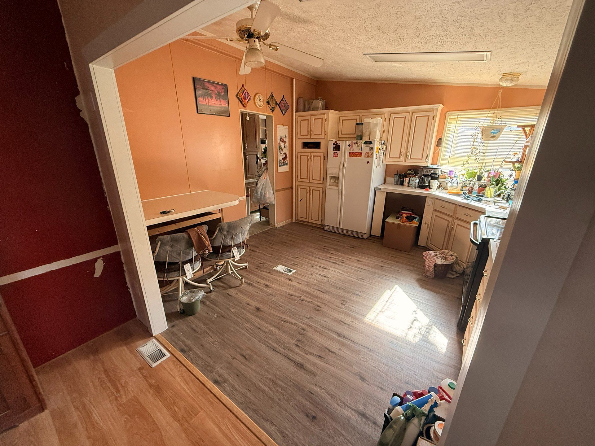 Small cluttered kitchen with peach walls, white cabinets, and sunlight by a window.