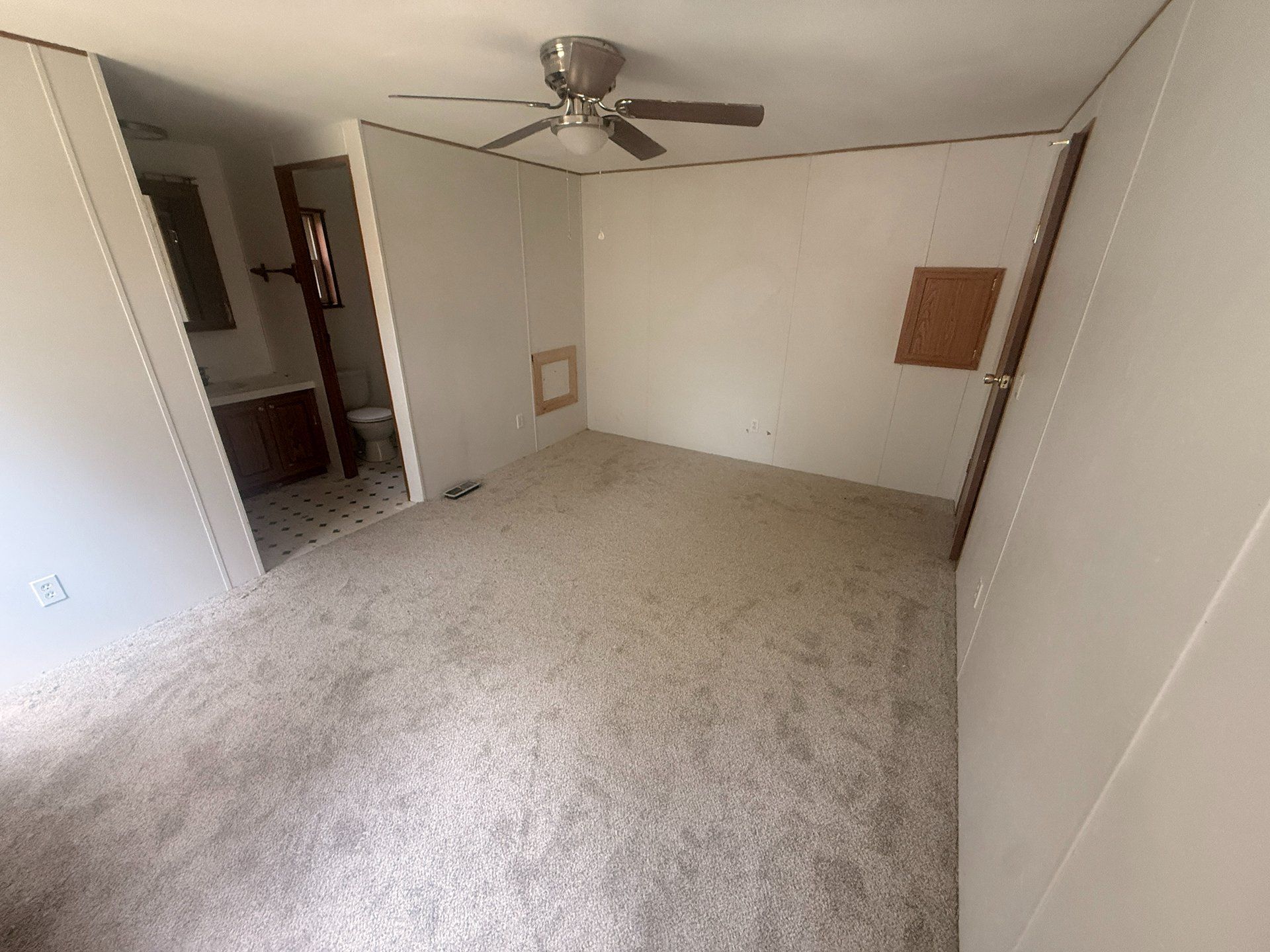 Empty beige carpeted bedroom with white walls, ceiling fan, and open doorway to a hall