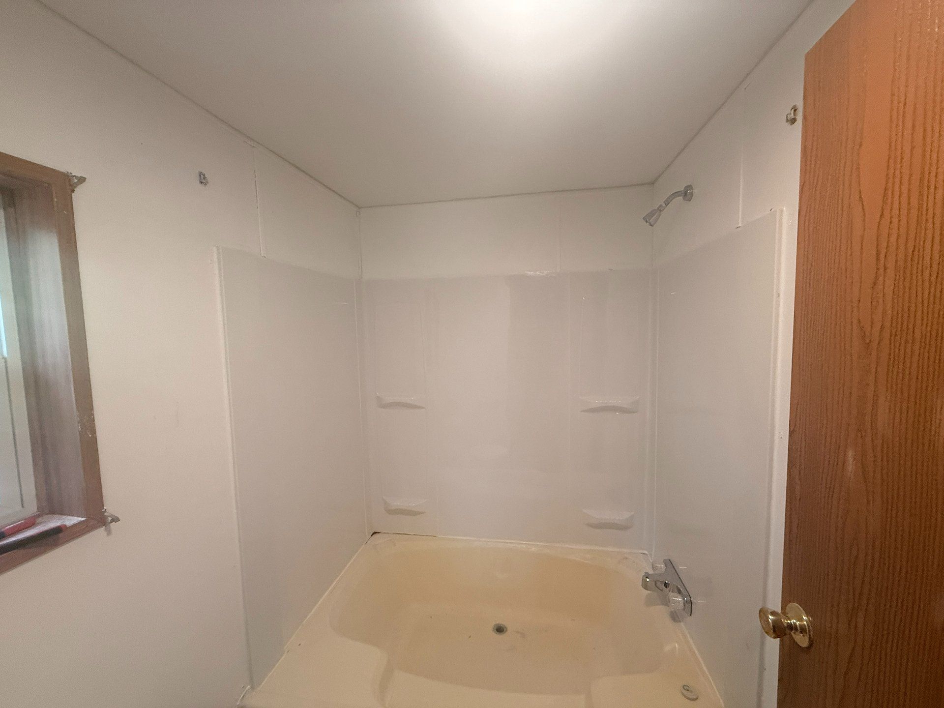 Small bathroom under renovation with white walls, beige tub, window, and open wooden door
