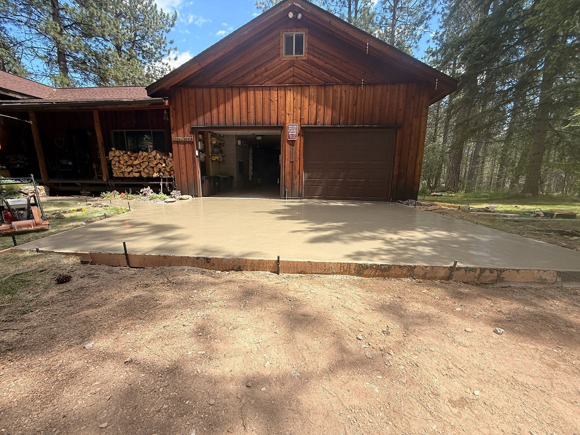 Rustic wooden garage and cabin with open center bay, concrete driveway, and pine trees in the background