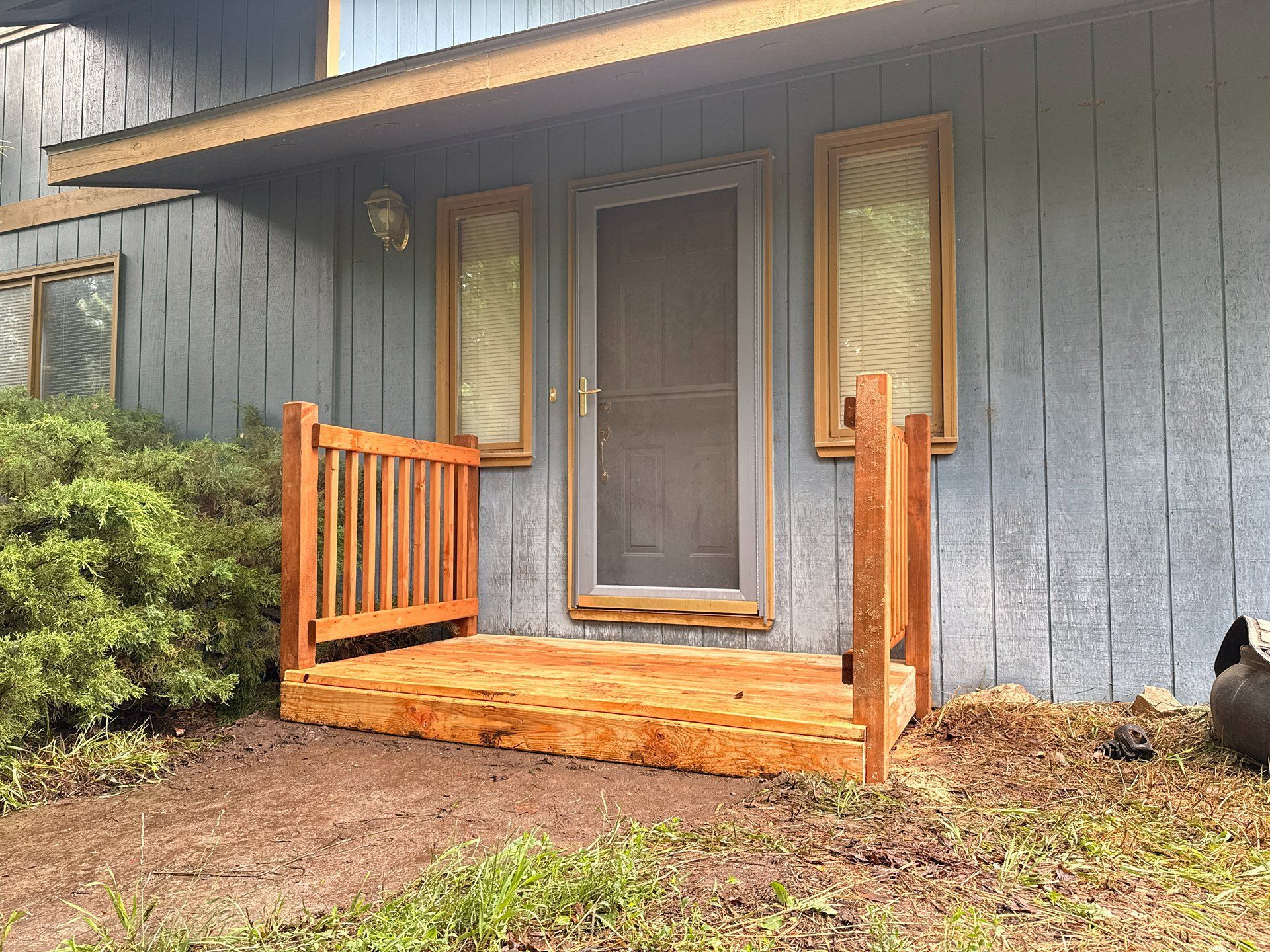 Blue house front porch with wooden railings and screen door, bordered by shrubs and dirt yard