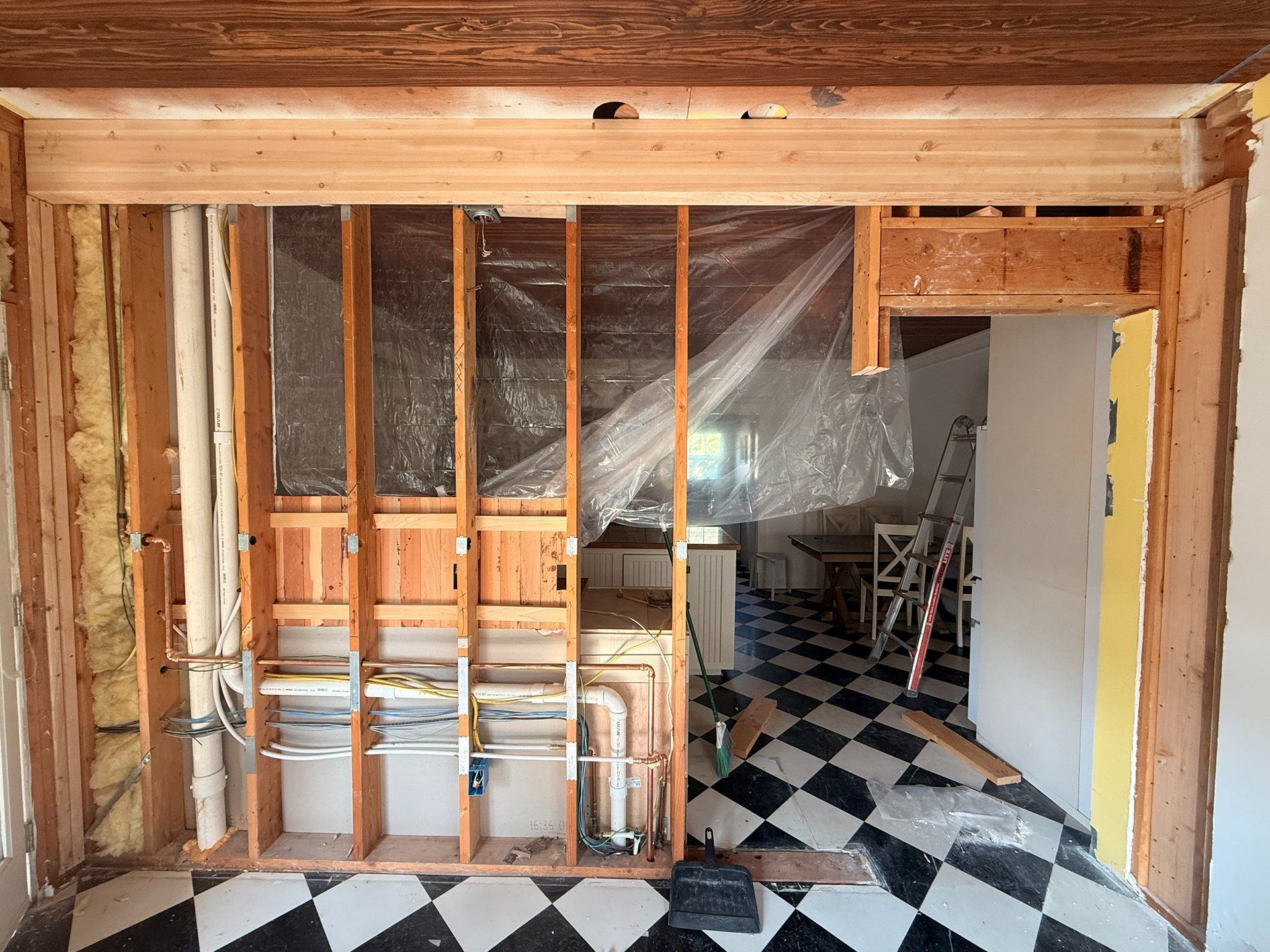 Unfinished room with exposed studs, plastic sheeting, and a black-and-white tiled floor leading into a doorway