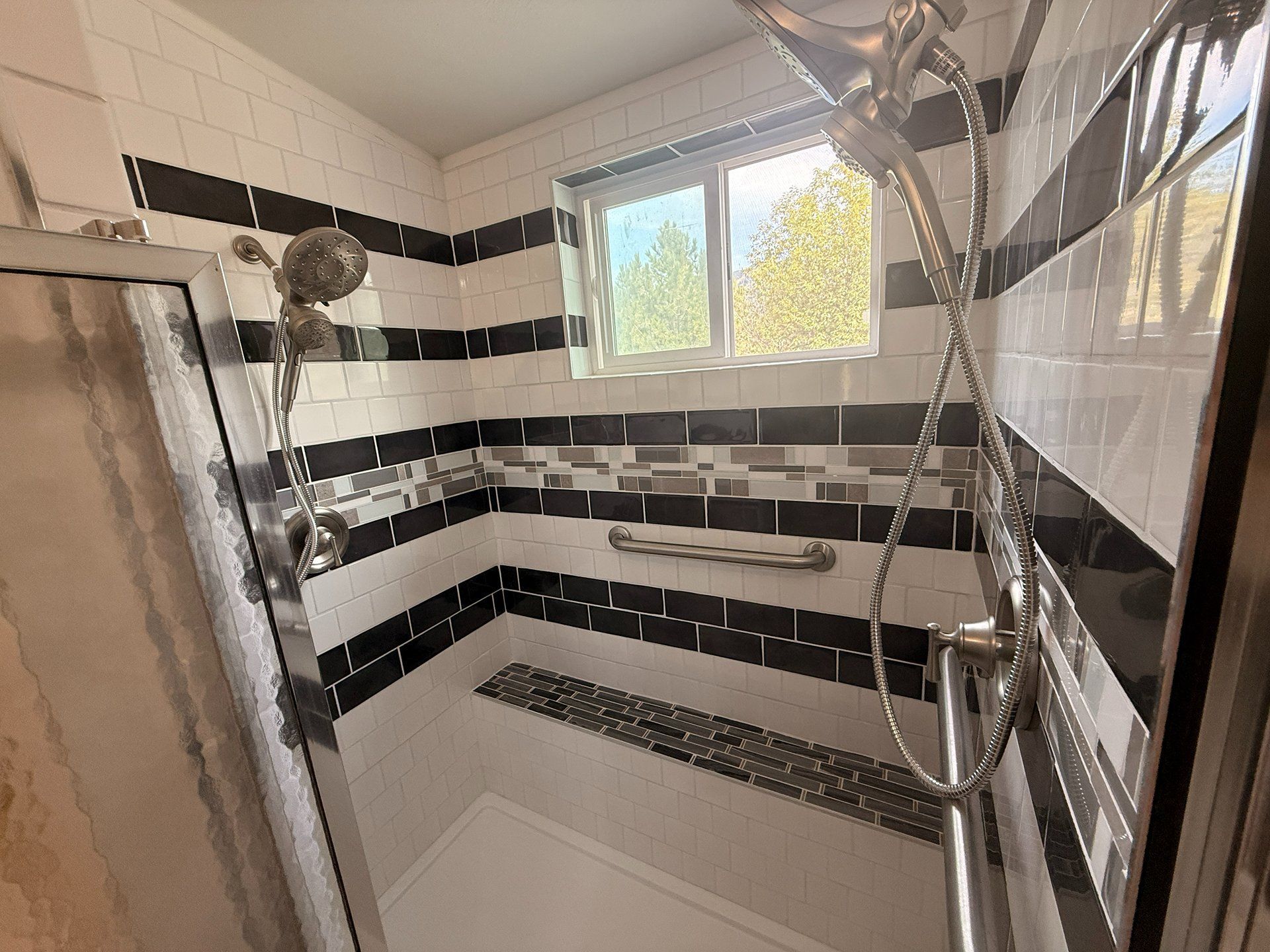 Bathroom with white and black tiled shower, handheld showerhead, and small window above the tub.