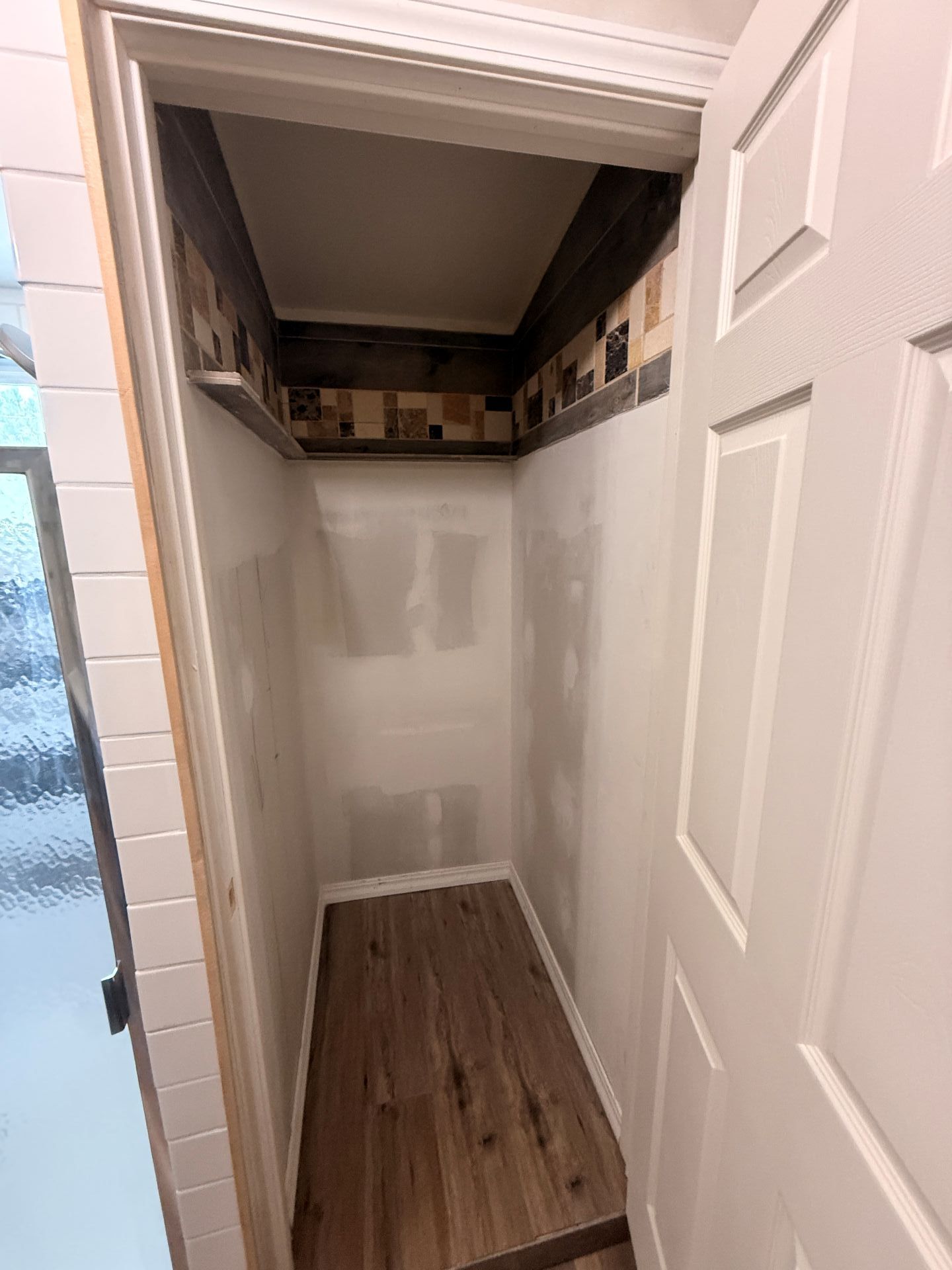 Small closet with open white doors, unfinished walls, and wood-look flooring; shelves line the top.