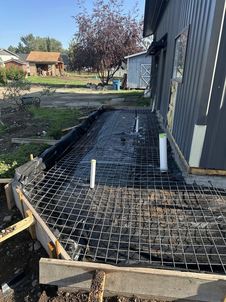 Concrete walkway under construction beside a dark gray house, with rebar grid and white PVC pipes exposed