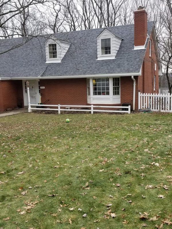 Brick house with gray roof, white picket fence, and dormer windows, in a grassy yard.