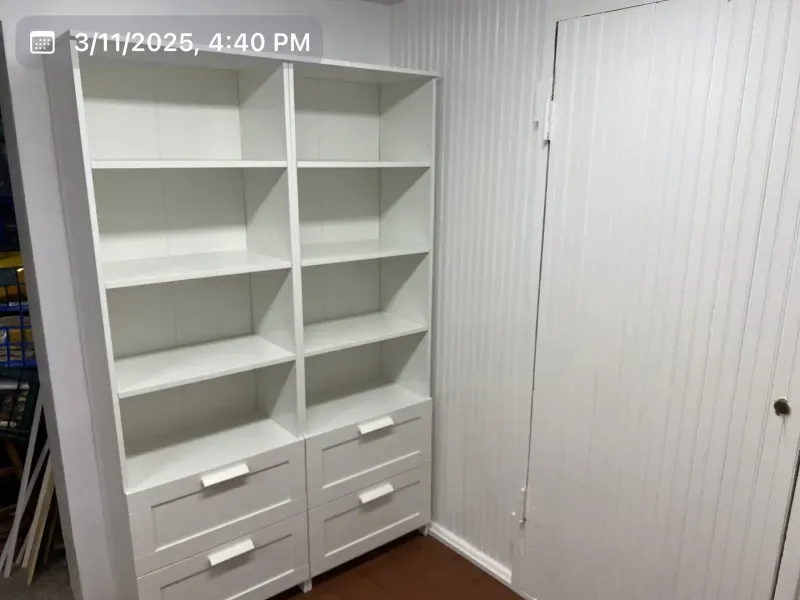 White bookshelf with drawers, against a white paneled wall and a closed white door.