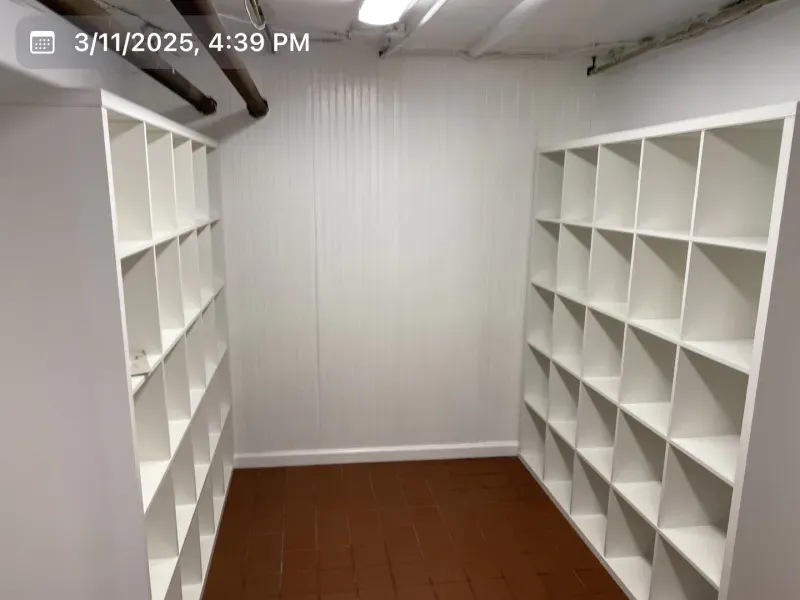 White shelving units line the walls of a small storage room with a brown floor and white walls.