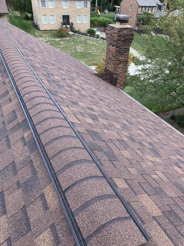 Brown asphalt shingle roof with a dark metal valley and brick chimney. Green grass and buildings in the background.
