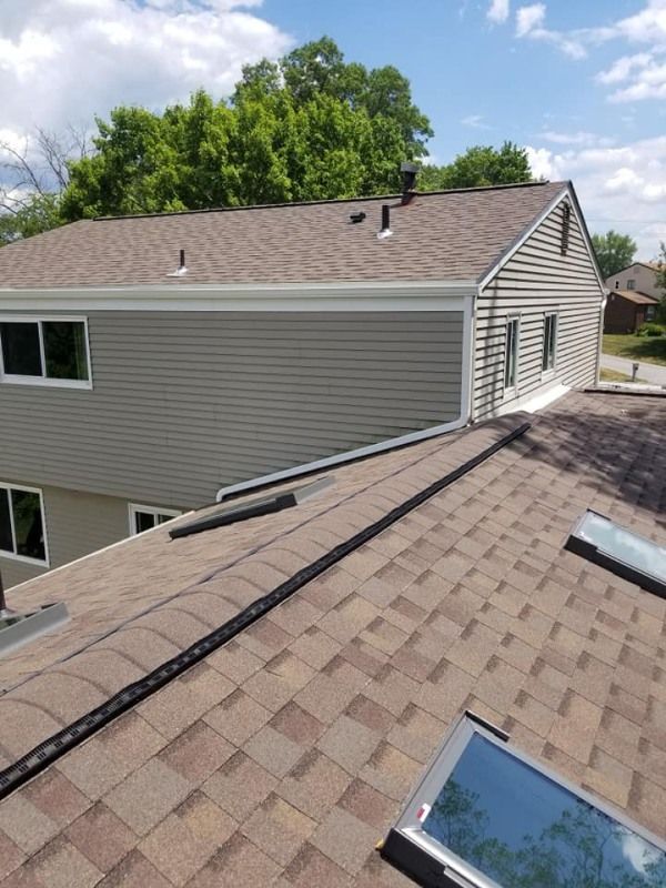 Brown asphalt shingle roof with black trim and a view of a gray-sided house with windows and trees.