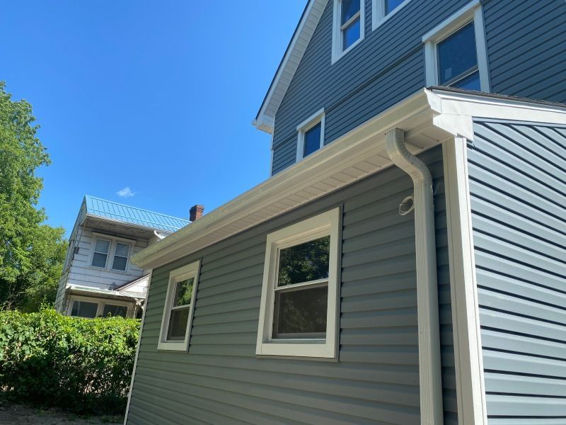 Blue-sided house with white trim and gutters, two windows visible. Clear blue sky in the background.
