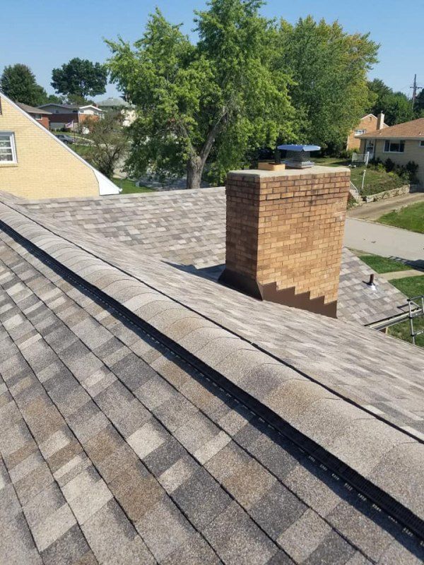 Roof with gray shingles, a brick chimney, and a tree in the background under a blue sky.