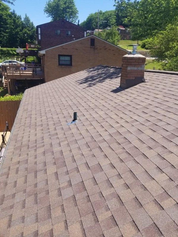 A brown shingled roof with a chimney and vent pipe. A house and trees are in the background.