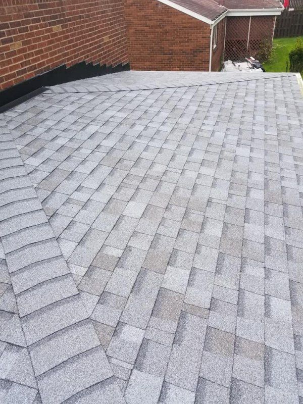 Gray asphalt shingle roof on a building, angled view. Brown brick wall and white siding in background.