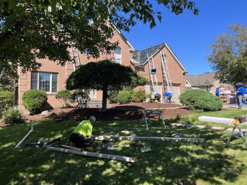 Workers install gutters on a brick house. Ladders and tools are on the roof and lawn; one worker in yellow vest.