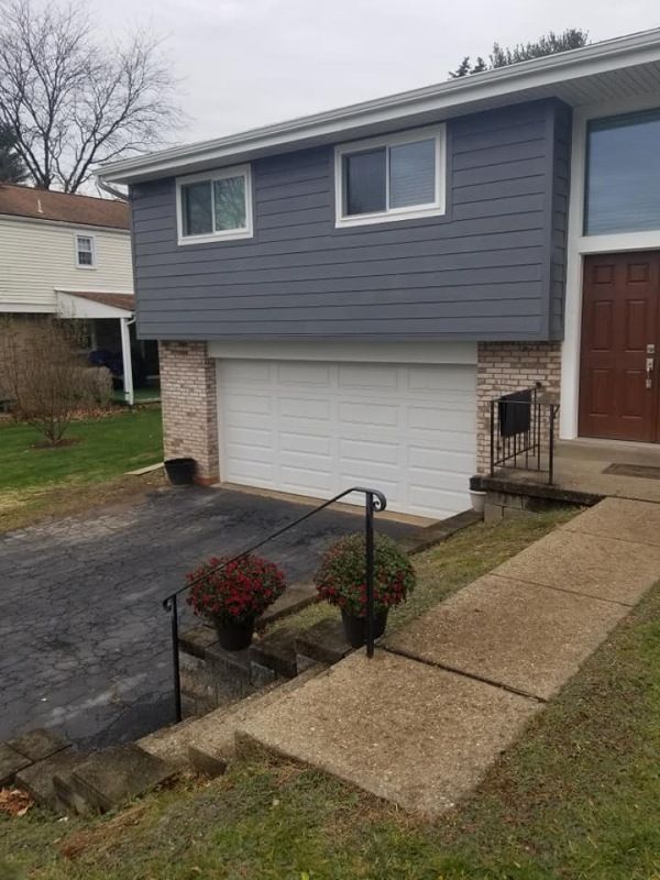 A two-story house with gray siding, a white garage door, and a brick facade. Black driveway and concrete steps.