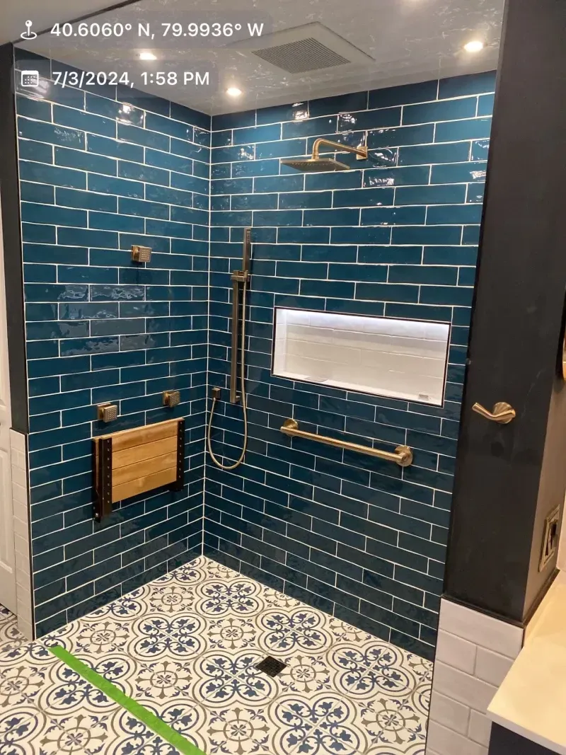 Blue-tiled shower with gold fixtures, decorative floor tiles, a built-in shelf, and a wooden seat.