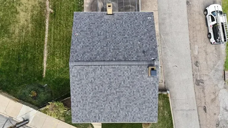 Aerial view of a gray asphalt shingle roof on a house, with a chimney and a white truck parked nearby.