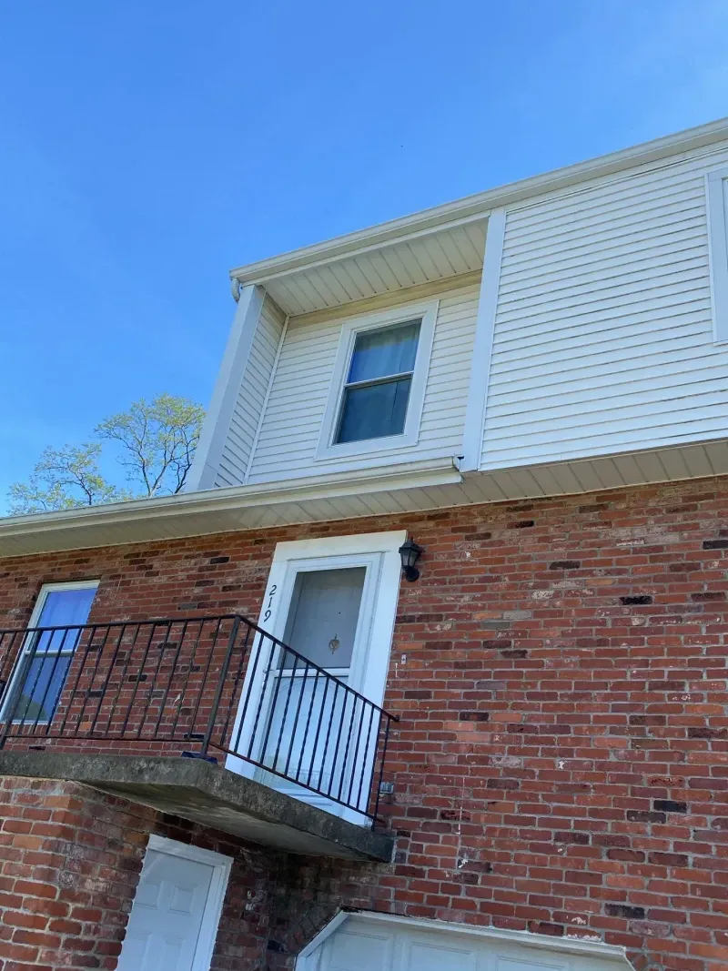 Red brick building with white siding, balcony, and dormer window against a blue sky.