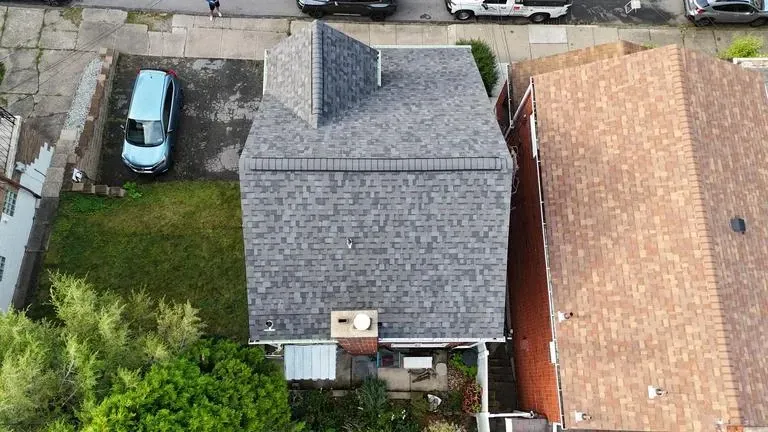 Aerial view of a gray-roofed house with a detached section, a blue car, and a brown-roofed neighboring house.
