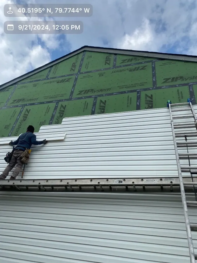 A worker installs white siding on a house under construction; a ladder leans against the wall.