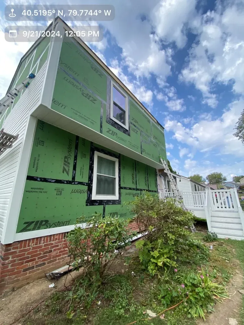 House exterior under construction, with green sheathing and white siding. Sunny, blue sky overhead.