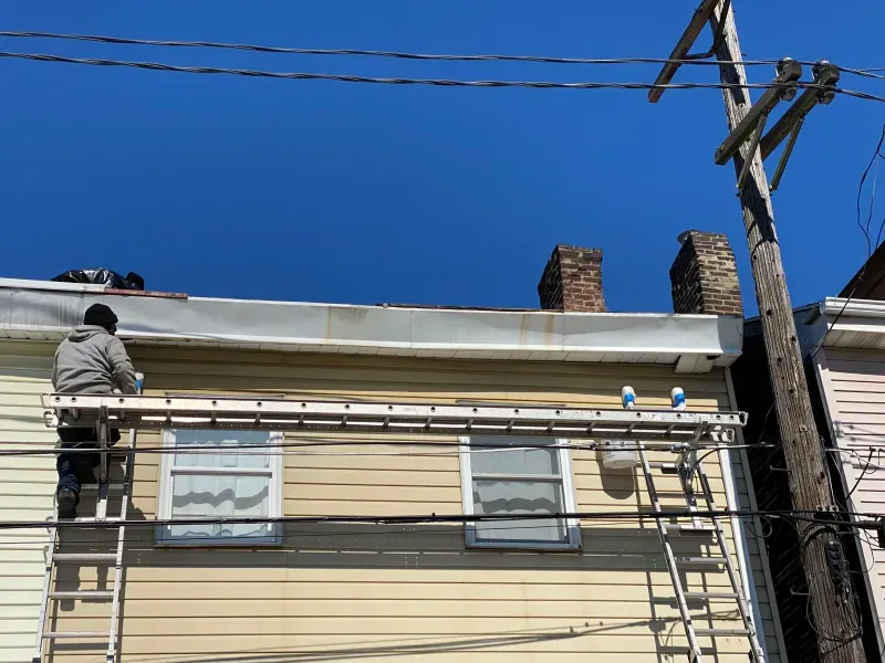 A person on a ladder repairs the roof of a yellow building with two chimneys under a blue sky.
