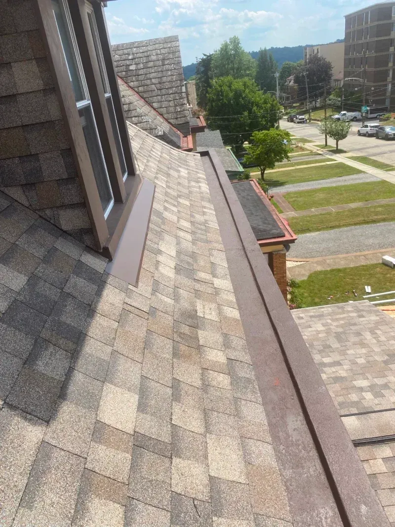 Roof of a building with brown shingles, metal flashing, and a window. Sunny day.