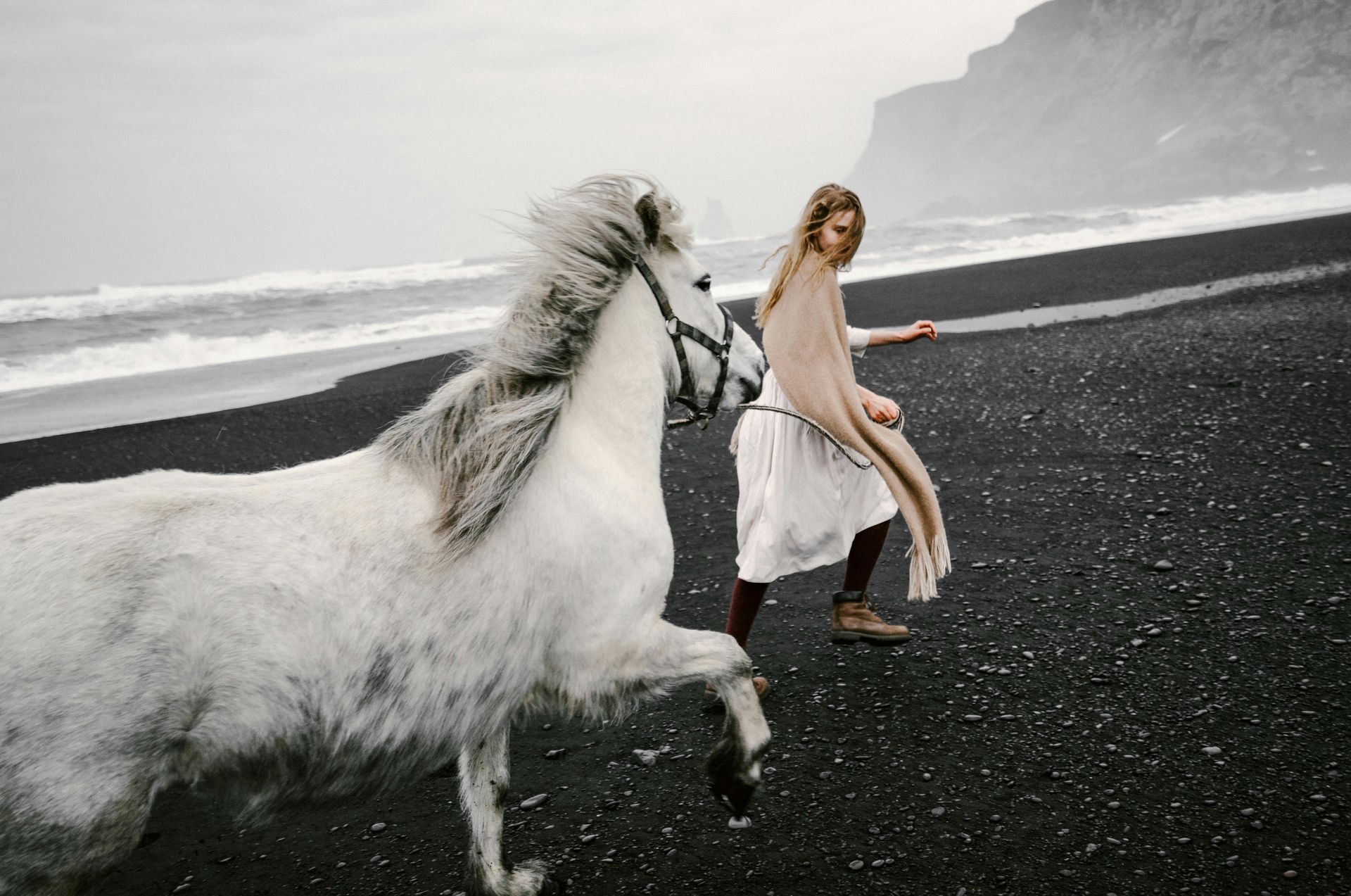 A woman is walking a white horse on a black sandy beach.